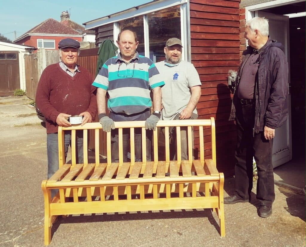 Four men standing behind a restored bench seat. Men in Sheds Lancing and Sompting